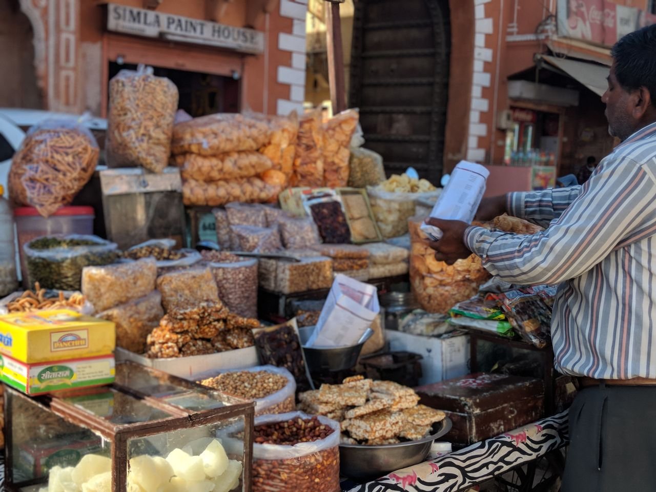 Vendor preparing a paper bag beside displays of snacks, nuts and sweets at a Jaipur market stall
